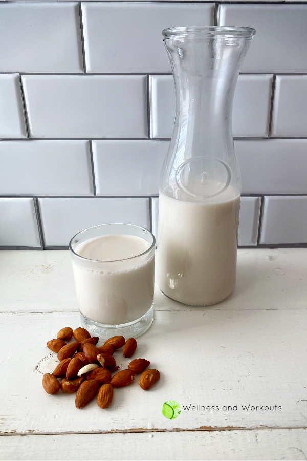 A glass carafe of delicious, fresh homemade Vitamix almond milk next to a glass of almond milk, with almonds in the foregroudn.