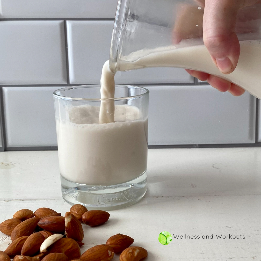 Side view of someone pouring homemade Vitamix almond milk from a glass carafe into a clear drinking glass, with dehydrated almonds in the foreground.