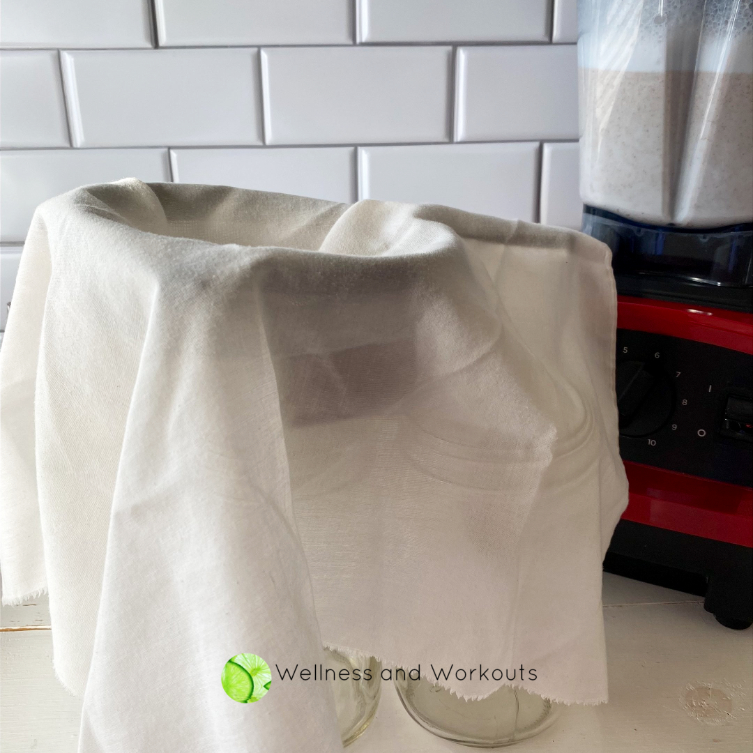 A square unbleached muslin cloth is draped over a wire strainer that sits on a canning funnel over a glass mason jar, ready to strain the Vitamix almond milk.